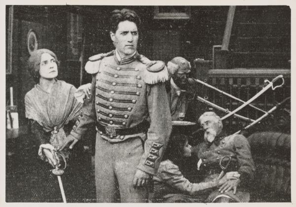 A scene from the 1910 film "A Dixie Mother" shows a woman (Florence Turner) holding a sword and looking at her son played by Carlyle Blackwell.  He is wearing a Confederate Army uniform.  An older man (Charles Kent) in uniform is sitting on a couch behind them and is being tended to by a young woman (Norma Talmadge) and a servant (probably played by a white man in blackface).