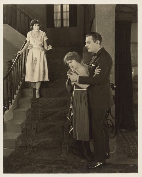 Norma Talmadge, playing Ann Hunniwell Regan, descends a flight of stairs and sees Frank Devereaux, played by Lew Cody, holding on a to a young woman (possibly Helen Weir as Helen Regan) in a scene from the 1921 film "The Sign on the Door".  Both women look upset.