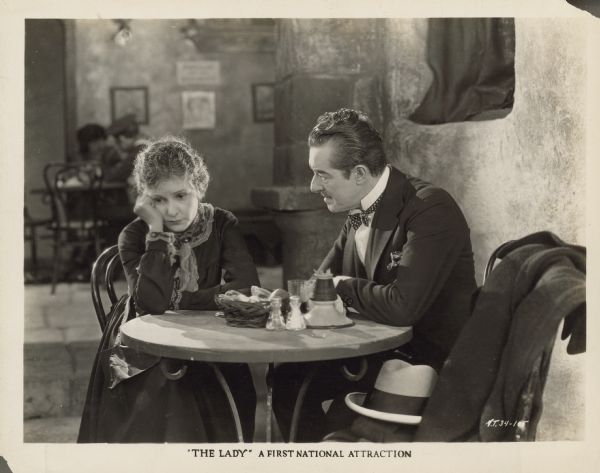 Norma Talmadge and Marc McDermott appear in a scene from the 1925 film "The Lady."  Talmadge, as Polly Pearl, sits at a table with her elbow on the table and her head resting on her hand - she looks very sad.  McDermott, as Mr. Wendover, sits next to her and looks at her.  They are sitting in a restaurant.