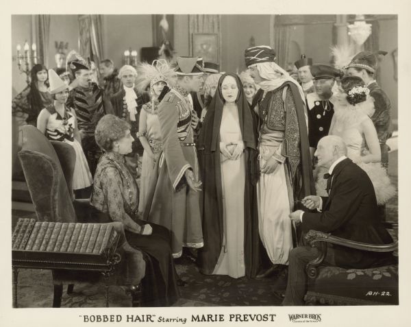 A scene from the 1925 film "Bobbed Hair" of people at a costume party.  Marie Prevost, as Connemara Moore, is dressed as a nun and stands between Reed Howes and John Roche - one wearing a military uniform and the other dressed as a sheik.  A crowd of people stand behind them and an older man and woman sit in chairs in front of them.
