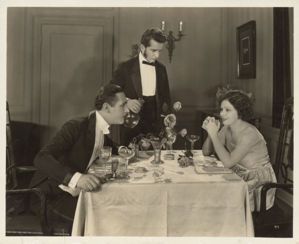 Norma Talmadge, as Ann Hunniwell, sits across a table from Lew Cody, as Frank Devereaux, in a scene from the 1921 film "The Sign on the Door".  The two are both dressed nicely and a waiter prepares to pour something from a pitcher.