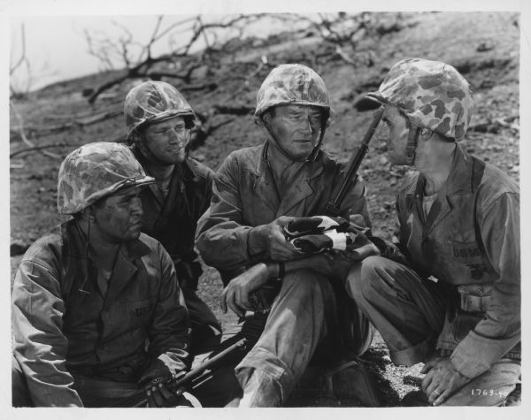 John Wayne and three other men sit on a hillside in a still from the 1949 film "Sands of Iwo Jima".  All are dressed in battle fatigues and Wayne holds a folded flag in one hand.  (see public note)