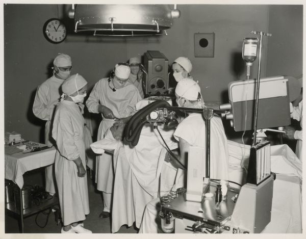 A doctor operates on a patient in a scene from the Ziv-TV Science Fiction Theatre episode entitled "Signals From the Moon".  Three women and one man dressed in  gowns, gloves, and masks, stand around the operating table.  Two large television cameras, one behind the table and one to the right, are present.