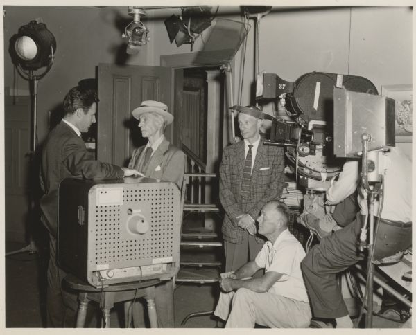 Peter Hansen talks to an older gentleman wearing a suit and a hat in a scene from "The Strange Lodger" episode of the Ziv-TV show Science Fiction Theatre.  They stand next to a television on a small table.  Three men, along with a film camera and lights, stand just out of the scene to the right.