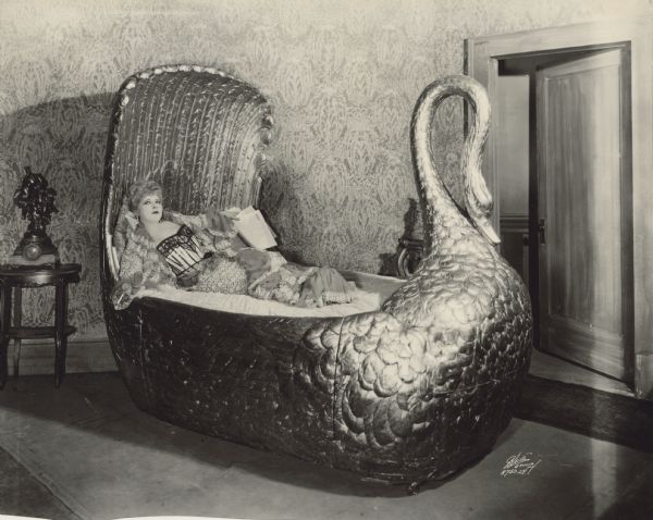 Publicity photograph of Mae West for the 1928 play <i>Diamond Lil</i>.  West lounges in a large bed shaped like a swan.  She holds papers in one hand a cigarette in the other.