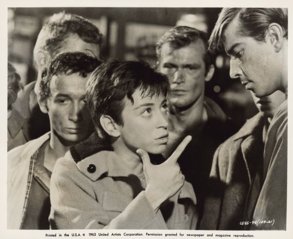 Publicity still for the 1961 film <i>West Side Story</i>. Anybodys (Susan Oakes) looks at a member of the Jets and holds their hand out like a gun. Four other members of the Jets stand nearby.  