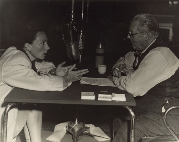 Agnes Moorehead sits across a small table and leans toward Lionel Barrymore.  Two large radio microphones hang in-between them.  The two were part of the radio series <i>Mayor of the Town</i>.  Barrymore holds a cigarette in one hand.
