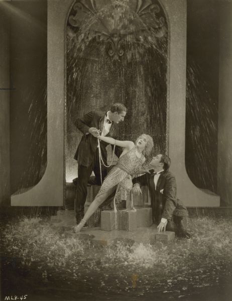 Circe (Mae Murray), Ballard Barrett (Charles Gerrard), and William Craig (William Haines) stand/sit underneath a water feature in a still from the 1924 film <i>Circe the Enchantress</i>.  
