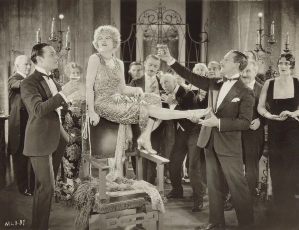 Circe (Mae Murray) sits on the back of a chair at fancy party.  She looks sideways at William Craig (William Haines) who stands next to her while Ballard Barrett (Charles Gerrard) holds a glass of champagne up towards her.  A crowd of people in tuxedos and gowns stand in the background and look on.