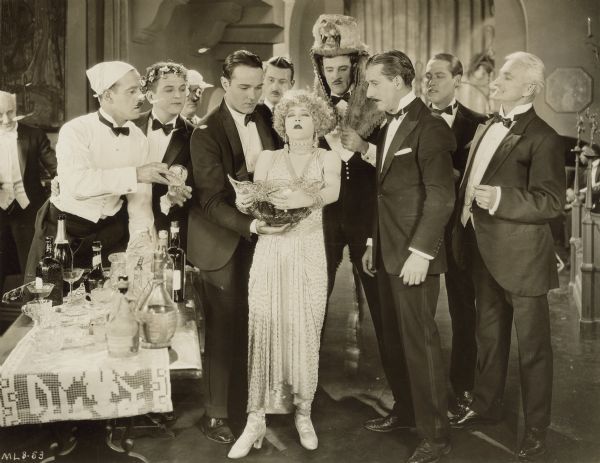 Circe (Mae Murray) stands with her eyes closed and holds a large punch bowl.  A group of men dressed in tuxedos stand around her - William Craig (William Haines) helps her hold the bowl while Ballard Barrett (Charles Gerrard) stands next to her.