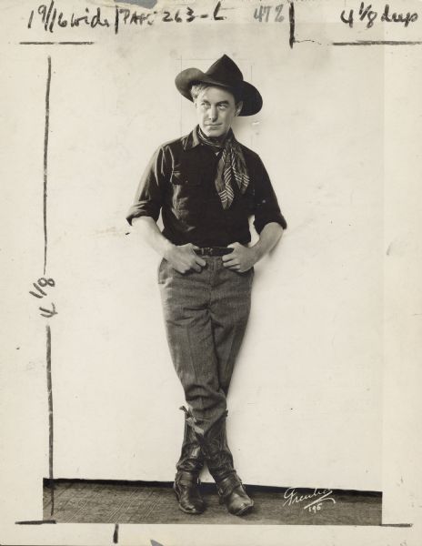 Publicity still of actor Harry Carey for the 1921 film <i>The Fox</i>.  Carey stands against a white wall and is wears a cowboy hat, neckerchief and cowboy boots.  His thumbs are hooked into his belt.