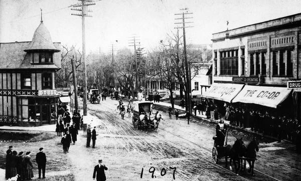 Elevated view of a costume parade on State Street, at the corner of State Street and Gilman Street. On both sides of the street are drugstores. The one on the right is the University Co-operative.