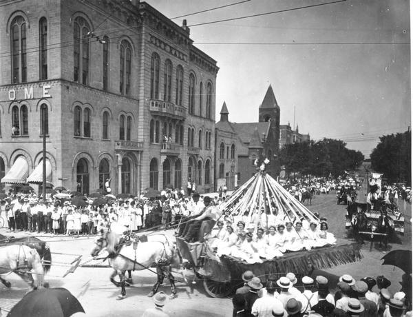 A parade turning the corner from Wisconsin Avenue on to the Capitol Square. A horse-drawn wagon in the foreground carries a May Pole and dancers from the Kehl Dance Studio.