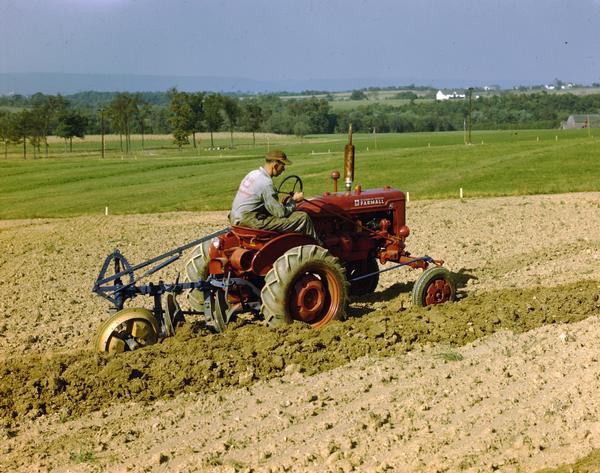 View towards a man driving a Farmall Super A tractor pulling a plow in a field.