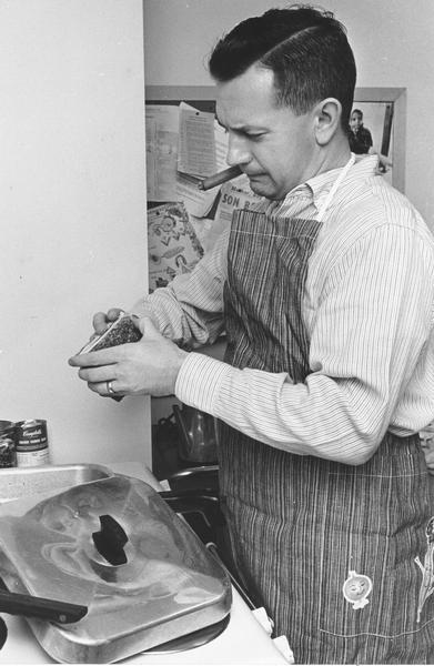 A father standing over a pan wearing an apron. He is cooking and smoking a cigar.