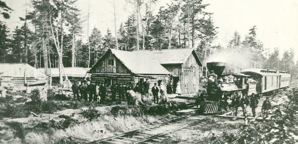 Slightly elevated group portrait of men and women gathered around the Turner House, which was erected by John Turner to house railroad men and construction officers. There are railroad tracks and a locomotive pulling railroad cars in the foreground.