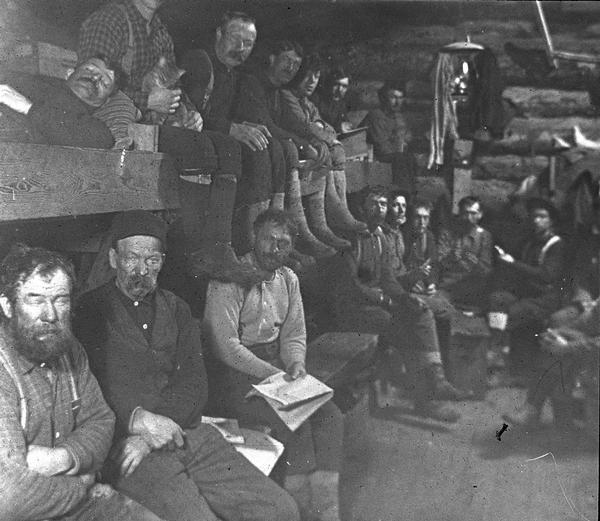 A logging crew poses inside their bunk house. In the upper left corner, one of the loggers holds a cat.