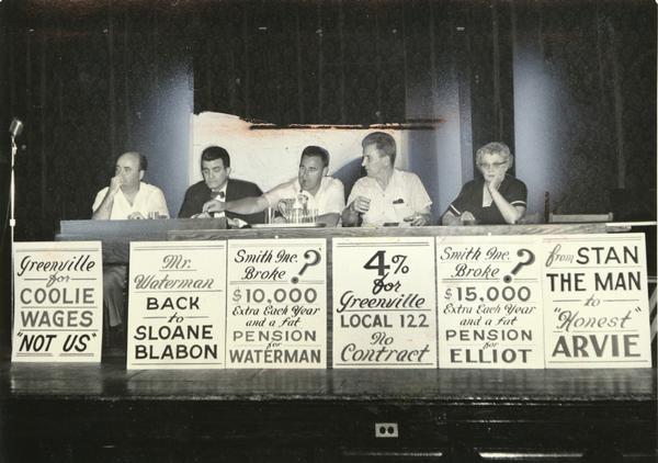 TWUA officials are shown seated at a table during a strike meeting with picket signs in front of the table. From left to right are business manager Charles E. Hughes, president Carmen Rose, 1st vice president John Kosorek, recording secretary John Hymes, and secretary-treasurer Mary Krupp.