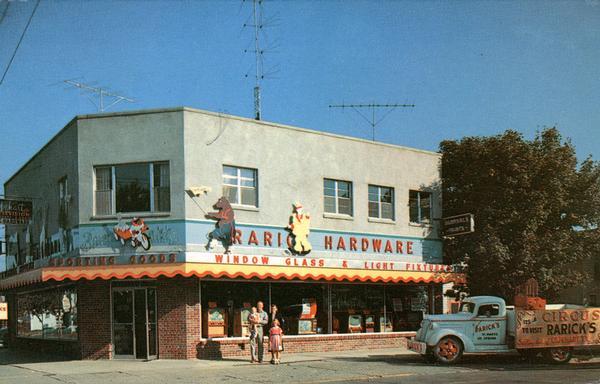 View across street toward Rarick's Hardware, a color television dealer. A group of people are posing on the sidewalk in front of the show windows. A truck is parked on the right.
