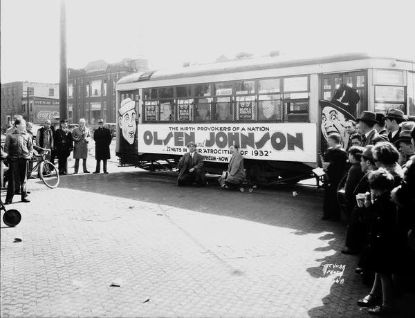 Comedians Ole Olsen and "Chic" Johnson, called "the mirth provokers of a nation," posing in front of a streetcar that displays an advertisement for their "Atrocities of 1932" performance at the Orpheum Theatre.
