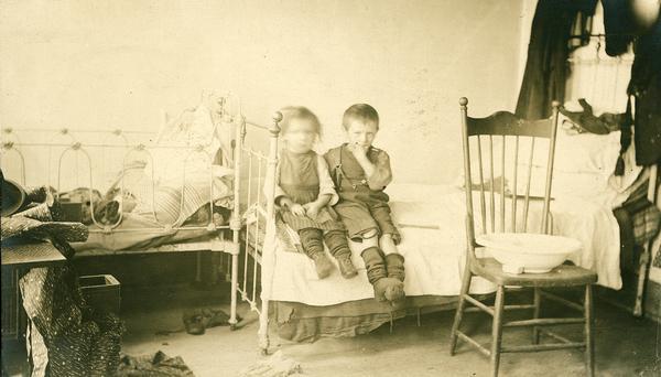 A young boy and girl sitting on an iron bedstead in the corner of their unkempt bedroom.