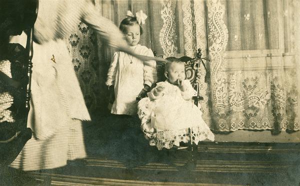 Interior scene of a small child in a rocking chair.  Standing next to the chair is a young girl.