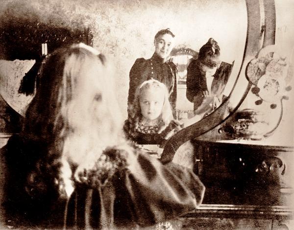 A woman photographer catches reflections of herself, a young girl and a woman in a bedroom bureau mirror.