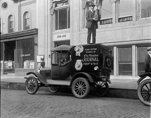 Standing atop a special vehicle fitted with a body built to resemble a camera, Taylor found the ideal vantage point from which to cover a news event. The car was equipped with a complete photographic darkroom that enabled him to develop his images on site. The side of the vehicle says: "News Picture Service of The Milwaukee Journal First - by Merit".