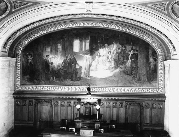 Interior of the Assembly Chamber in the Wisconsin State Capitol showing the mural painted by Edwin H. Blashfild. The painting depicts the "State of Wisconsin; its Past, Present, and Future." "Wisconsin" is seated in the center, surrounded by three female figures representing Lake Superior, Lake Michigan, and the Mississippi River, and various historic figures from her past. The standing figure in the center represents "Today," pointing to the accomplishments of Wisconsinites in the distance. Behind her stand miners, farmers, and lumbermen with their families. At the far left, "Future" shelters her "Lamp of Progress" and listens to the spirit of conservation. 
The mounted eagle behind the Speaker's chair is a bald eagle, the same type as "Old Abe," a famous Wisconsin Civil War mascot. 
