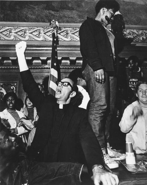 Father James Groppi with his fist in the air at the Wisconsin State Capitol during welfare demonstrations.He is surrounded by other protesters.