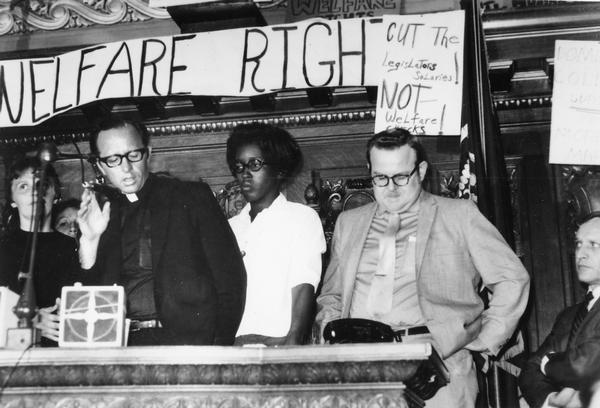 Father James Groppi speaking at a demonstration at the Wisconsin State Capitol Assembly Chamber protesting welfare cuts. Father Groppi is seated on the left. Rep. Harold Froehlich is on the right.