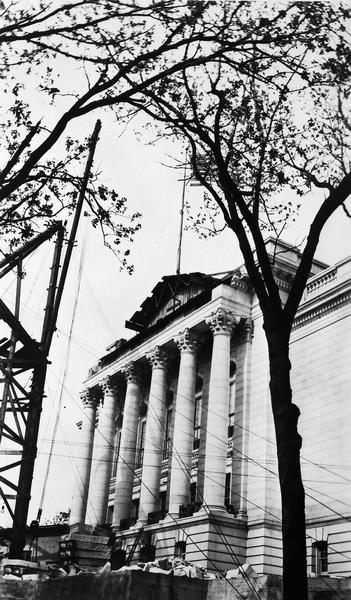 The west wing of the third Wisconsin State Capitol on Decoration Day. The steep slope of the western side of the park delayed the construction of the east portico and pediment until dirt from the East Wing terrace could be used to build up the terrace and allow the Woodbury Granite Company to set up its derricks. During construction, the partially completed pediment collapsed, killing one setter.