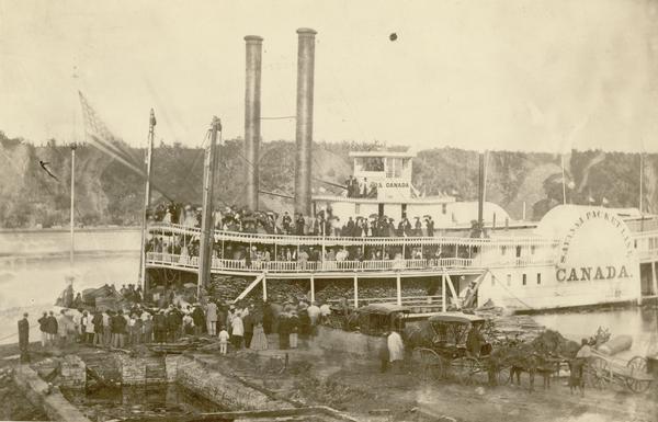 The steamboat "Canada" at dock; cargo on lower deck, passengers on upper deck, and crowd waiting on the dock. The "Canada" was built in 1858, dismantled in 1870, and operated on the upper Mississippi.
