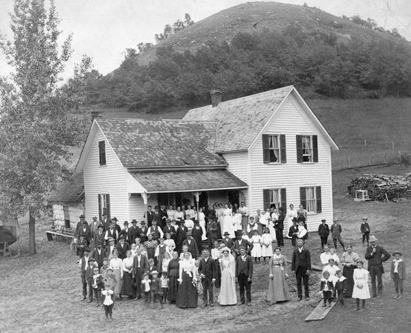 People attending a wedding reception, posing outdoors. They are standing in front of a modern house that has a log cabin attached to the back. In the background is a round, tall hill.