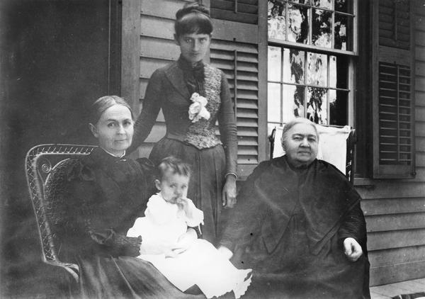 Three women and a baby girl posing on a porch outside a house. Seated on the left is Louise Sophia Favill, standing is Eleanor Favill Tenney, seated on right is Elizabeth Therese Baird. The baby is Elizabeth Louise Tenney.