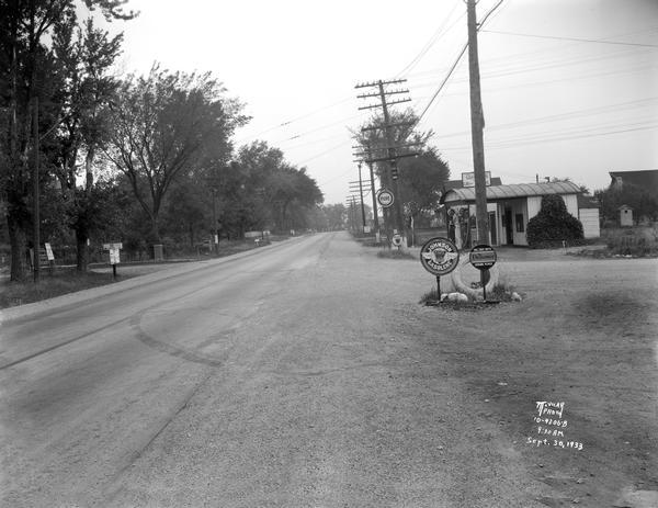 A gas station in a Trachte building on the corner of Highway 51 (Monona Drive) and Highway 30 (Cottage Grove Road). "Johnson Gasoline," and "Defiance Sparkplugs" are advertised. The John W. Walterscheit Jr., farm is in the background behind the gas station.