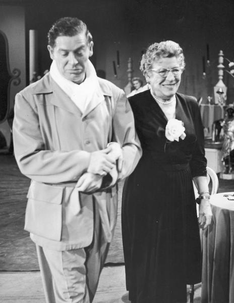 Milton Berle, dressed in a suit with a white neckscarf, is shown escorting his mother Sandra.