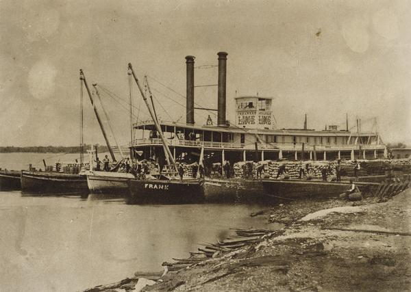 The sternwheel packet, "Arkansas," loaded with gunny sacks of wheat taken between 1872 and 1878. Men are unloading barrels and gunny sacks on a gangplank that crosses some of the wharf barges that are alongside her.