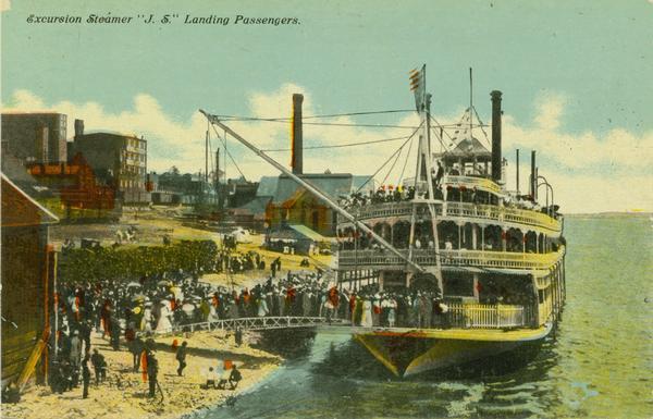 The sternwheel excursion, <i>J.S.</i>, landing passengers, taken between 1901 and 1910. Crowds are on riverbank and there are buildings in background. A crane on the front of the steamer is attached to a passenger boarding bridge.