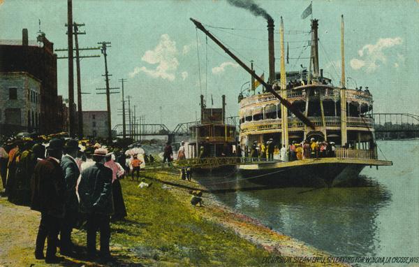 View along shoreline of the sternwheel excursion, <i>J.S.</i>, getting ready to leave for Winona. A crowd is watching from the landing. A crane on the front of the steamer is attached to a passenger boarding bridge.