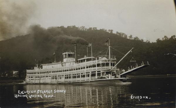 The sternwheel excursion, <i>Sidney</i>, leaving Lansing, Iowa. Later named <i>Washington</i>. A crane on the front of the steamer is attached to a passenger boarding bridge.