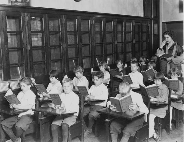A group of young students seated in their classroom holding books as the teacher stands at the back of the room. The title of the book is "The Progressive Music Series, Book One" and the teacher is holding a harmonica up to her mouth.