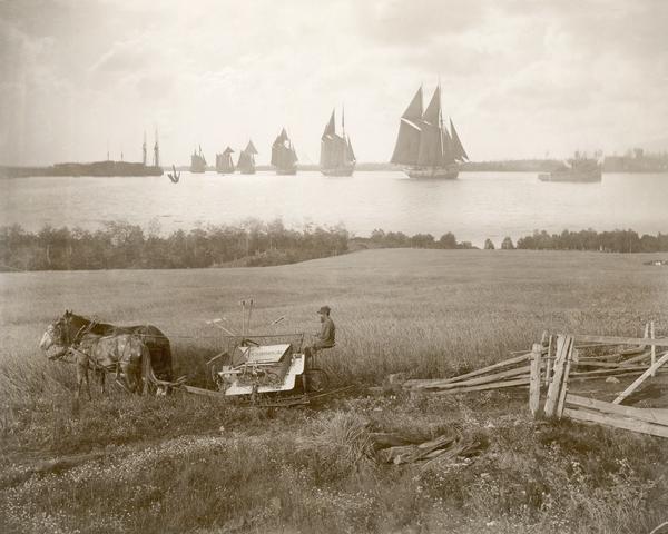 Man operating a McCormick horse-drawn grain binder in a field. A tugboat is pulling sailing ships on a body of water in the distance. The photograph appears to be a composite of two different images.