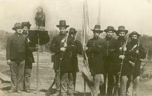 "Old Abe" and "Color Guard" of the Eighth Regiment, Wisconsin Volunteer Infantry, posed in uniform, outside with Old Abe on a shield perch. Likely Edward Homaston of Eau Claire holding the perch. Sgt. Ambrose Armitage is third from left. The portion of the photograph featuring Old Abe appears to be altered.