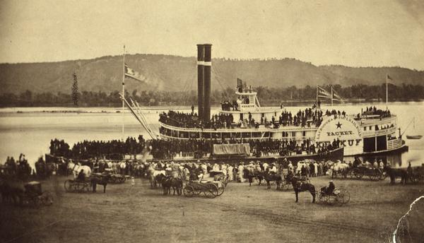 The sidewheel packet, <i>Keokuk</i>, loaded with passengers beside a barge at a landing. The landing is filled with people and horse-drawn vehicles. The sign on the side of the wheel reads: "La Crosse and St. Paul Railroad, Packet, Keokuk." Bluffs are on the opposite side of the river.