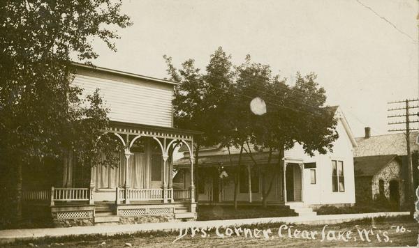 Photographic postcard of across street toward buildings, which include the offices of Dr. L. Campbell (house on the left) and Dr. Anton Nelson, the father of Gaylord Nelson. Caption reads: "'Doctors' Corner,' Clear Lake, Wis."