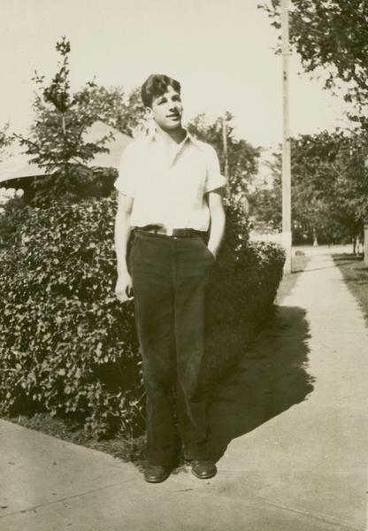 Gaylord Nelson during his high school years wearing a white, short-sleeved shirt standing in front of hedges and a bandstand.