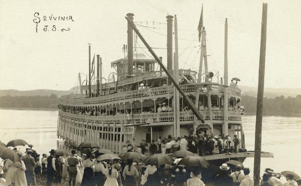 The sternwheel excursion boat, <i>J.S.</i>, at a landing at Cassville. People with umbrellas wait on shore. Inscribed <i>Souvenir J.S.</i>  The <i>J.S.</i> was a successful excursion boat. She was destroyed by fire at Victory, Wisconsin, June 25, 1910, and it was said that the fire was set by a passenger who was confined in the jail in the hold of the boat.