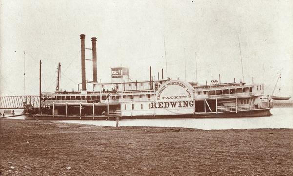 The sternwheel packet, <i>Red Wing</i>, at a landing on the upper Mississippi River between 1870 and 1882. There is a bridge behind the boat on the left. Sign painted on side of boat reads: "Keokuk Northern Line Packet Redwing."