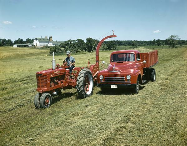 Man driving an International Harvester Farmall M tractor which is pulling a forage harvester next to a truck.